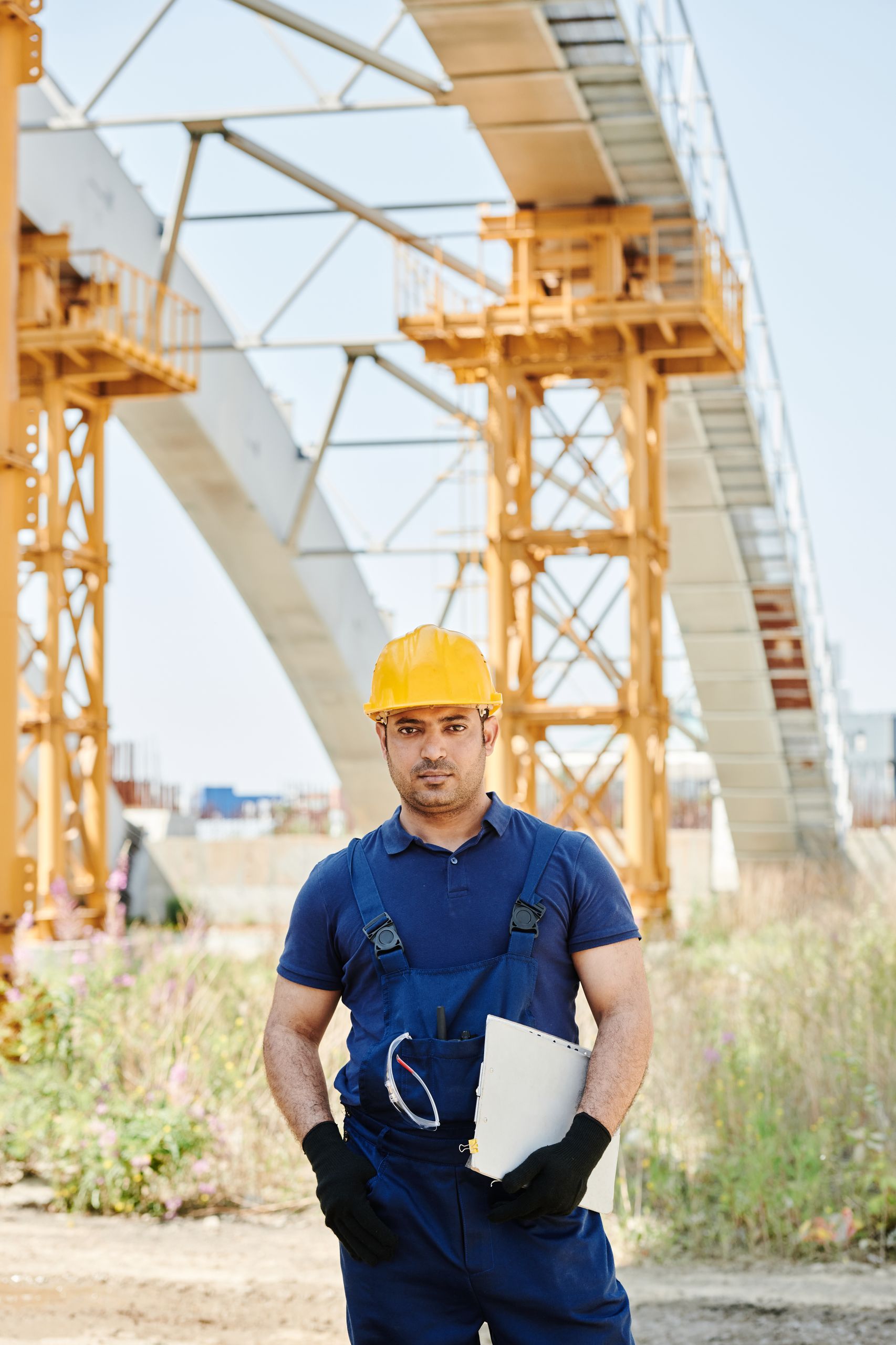 supervisor in safety gear holding a clipboard at a construction site with arches in the background.